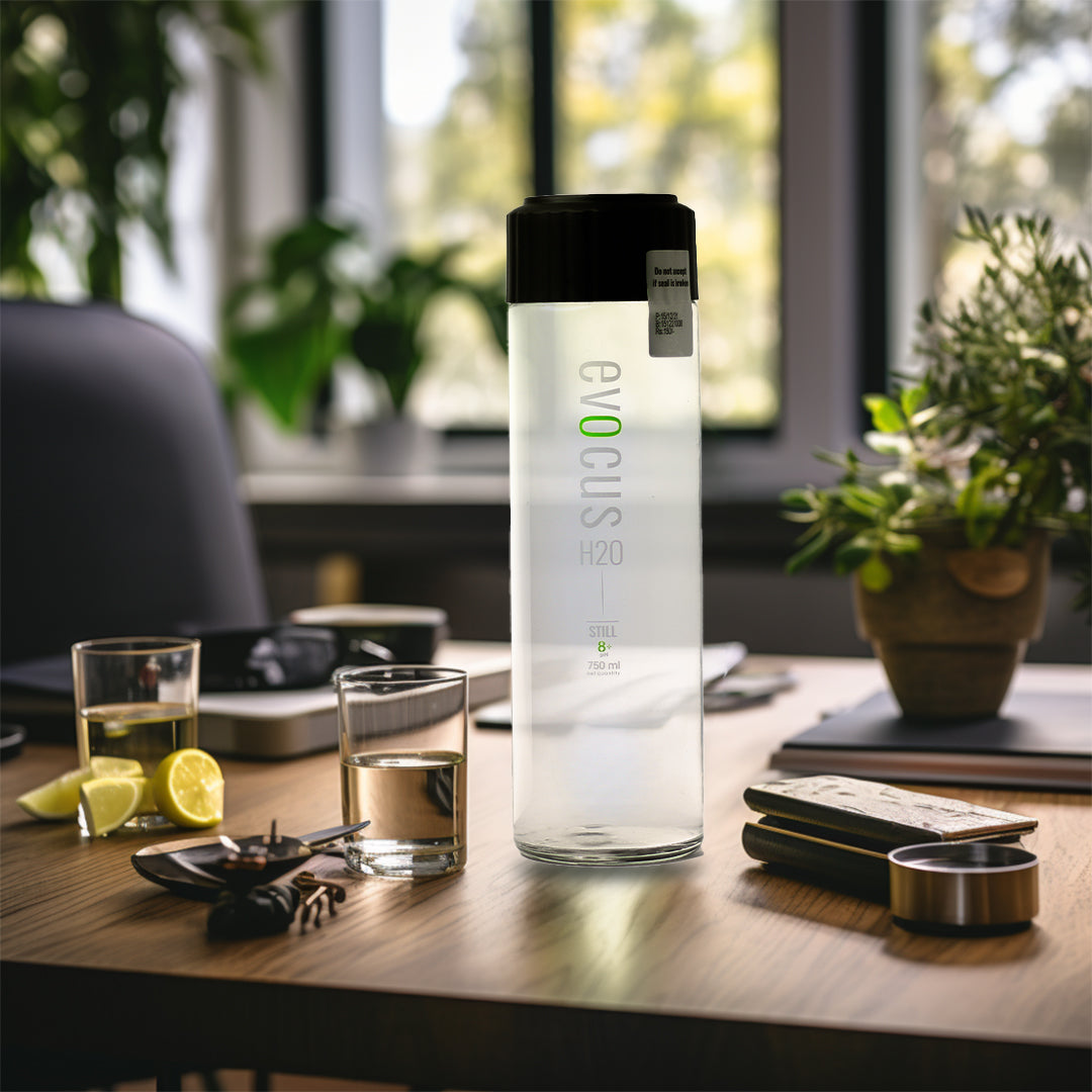 A 750ml glass bottle of Evocus Alkaline Water sits on a wooden desk in a home office setting.