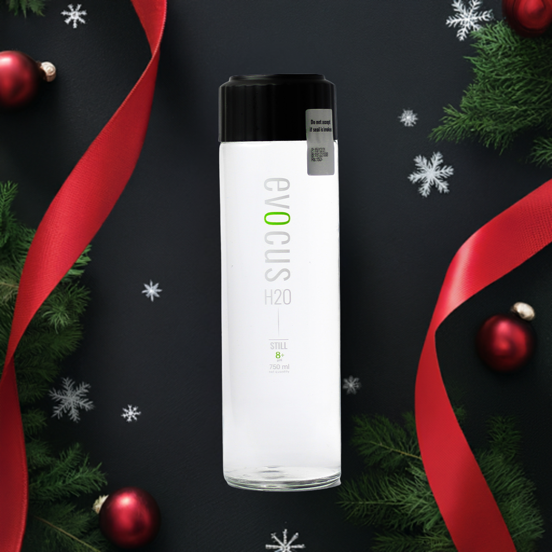 A 750ml glass bottle of Evocus Alkaline Water sits on a dark background surrounded by festive Christmas decorations like red ribbons and ornaments.